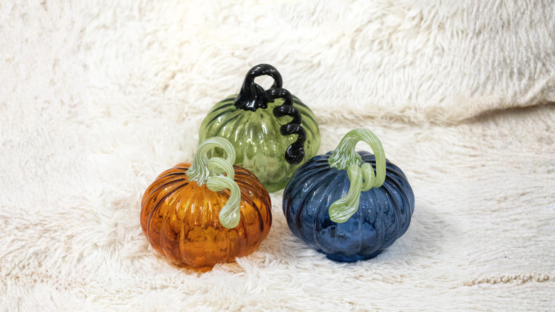 Three glass pumpkins in orange, green, and blue on a textured white surface